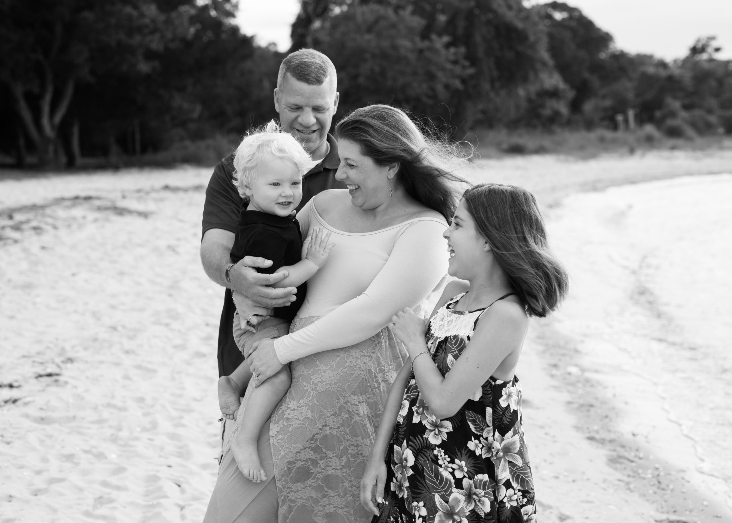 Pregnant mom at Yorktown beach hugging her toddler with her family surrounding her, getting her maternity pictures done by a Yorktown maternity photographer.